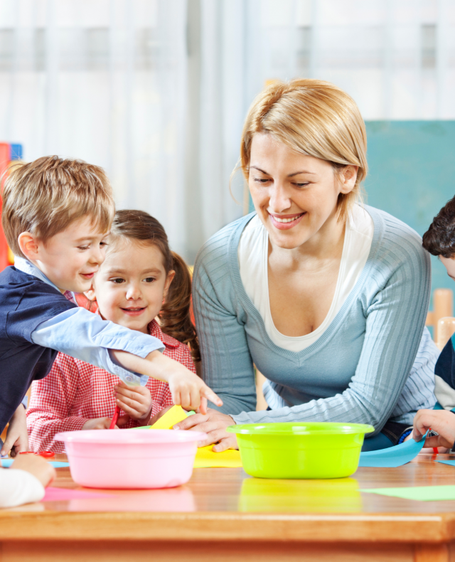 Afbeelding van juf die met 4 kinderen aan een tafel zit met 2 bakjes en gekleurd papier